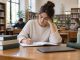 Persona joven estudiando en una biblioteca con cuaderno, libros y ordenador cerrado
